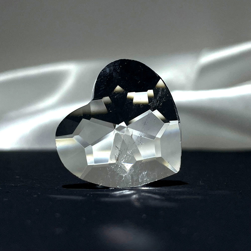 A faceted clear quartz heart dramatically lit from the side on a dark surface.
