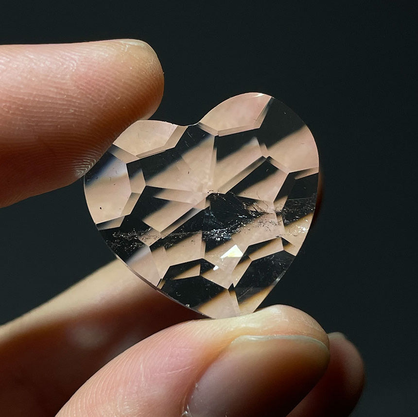 Fingers holding a small, faceted clear quartz heart up to the light to show its clarity