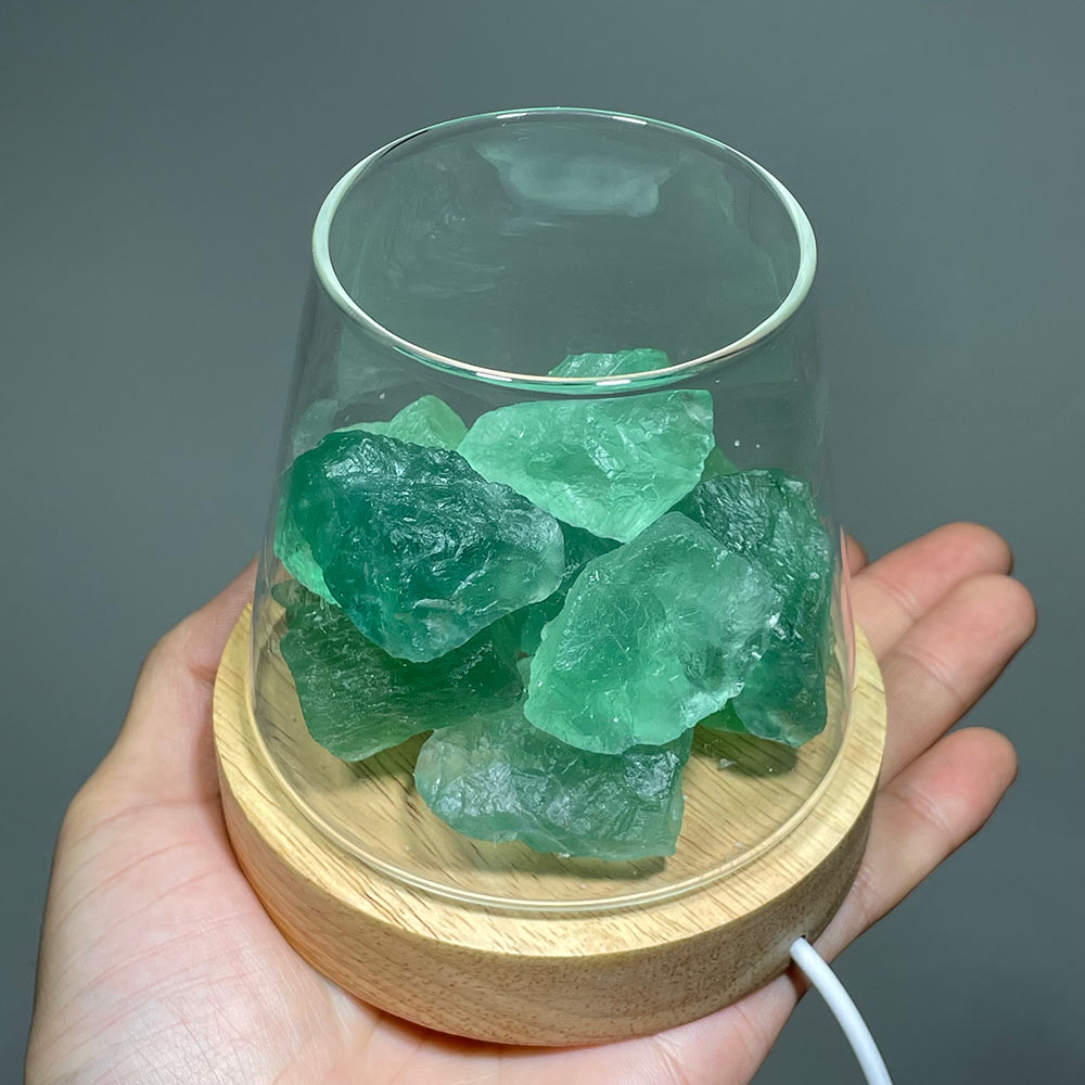 Hand holding a glass lamp filled with raw green fluorite crystals on a wooden LED base