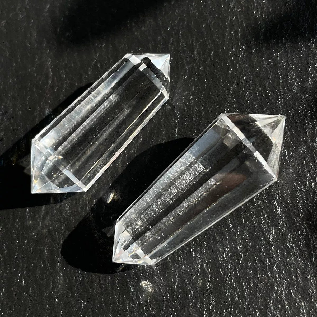 Two 12-sided clear quartz Vogel wands lying on a dark, textured surface.