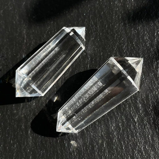 Two 12-sided clear quartz Vogel wands lying on a dark, textured surface.