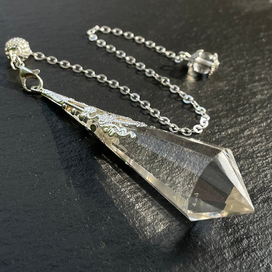 Faceted clear quartz pendulum with an ornate silver cap and chain on a black surface.