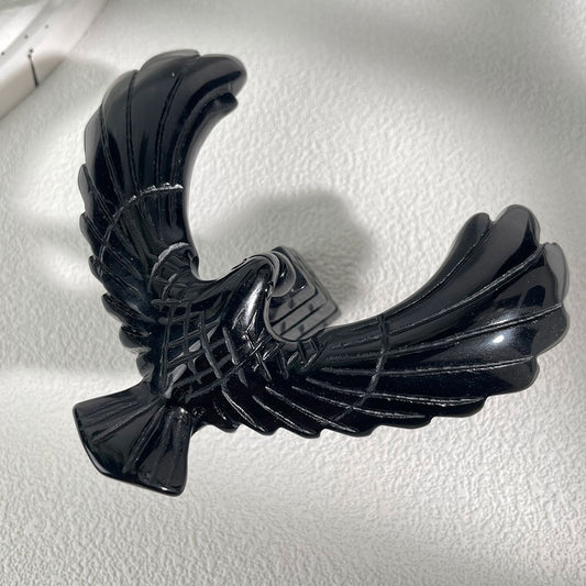 A glossy black obsidian eagle perfectly balancing on its beak atop a carved stone pyramid base against a floral background.