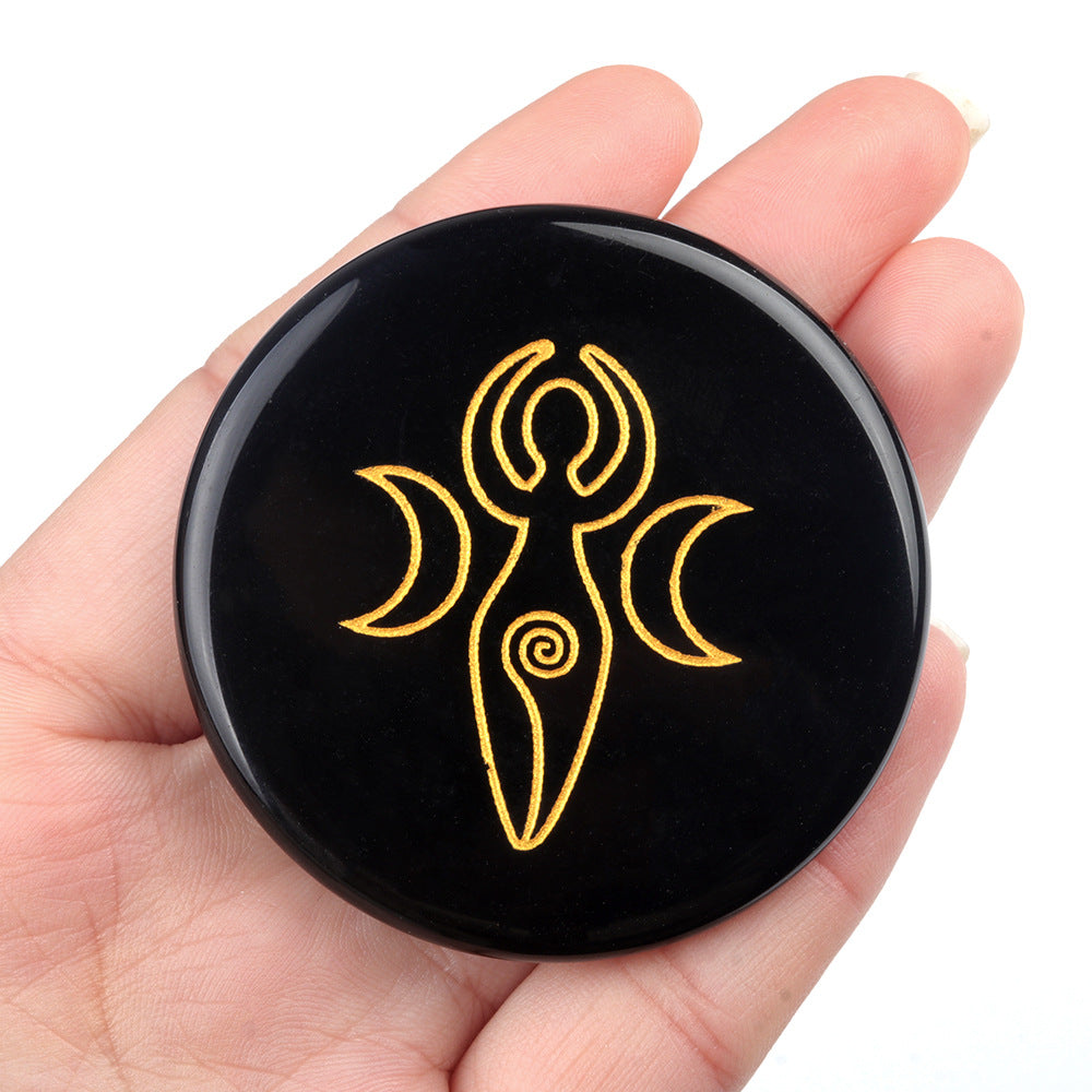 Hand holding a polished black obsidian disc engraved with a golden Spiral Goddess symbol.