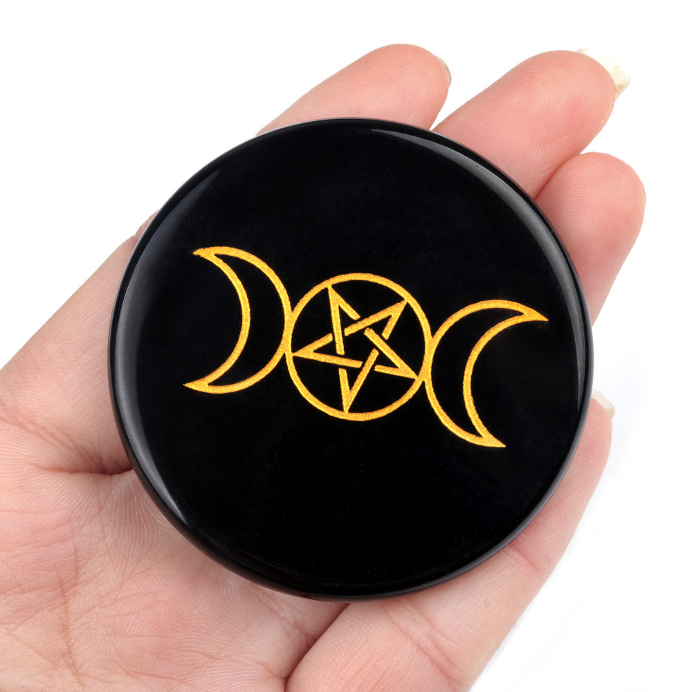 Black circular object with gold pentagram and moon symbols held in a hand against a white background
