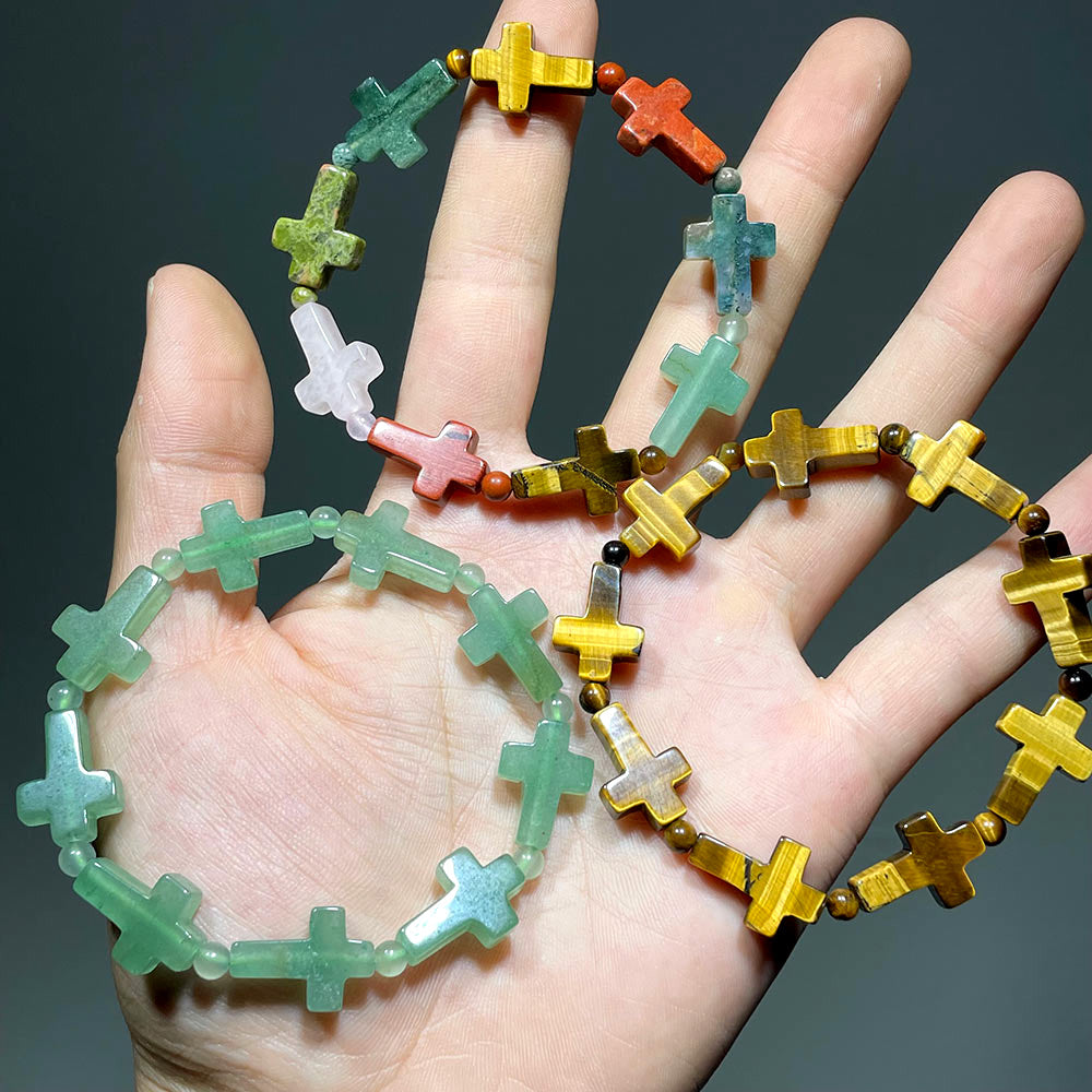 Hand holding three crystal cross bracelets in Green Aventurine, Tiger's Eye, and a mixed gemstone version