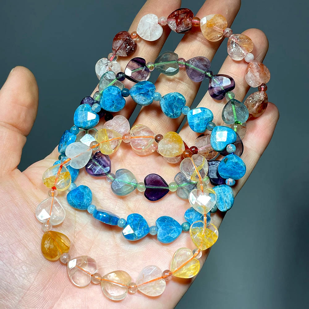Hand holding a cascade of mixed gemstone faceted heart bracelets, including fluorite, apatite, and fire quartz
