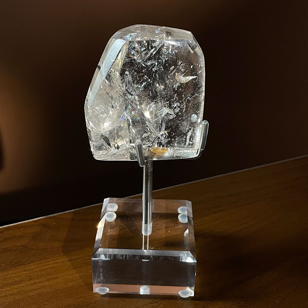 A highly transparent natural Madagascar quartz polished freeform crystal, shaped like a shield, resting on a clear acrylic stand.