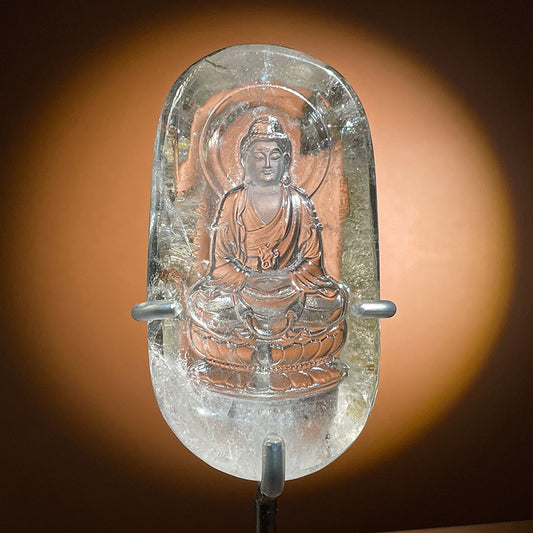 A highly transparent natural clear quartz crystal plaque featuring a frosted, deep-relief carving of a meditating Buddha, resting on a clear acrylic stand.