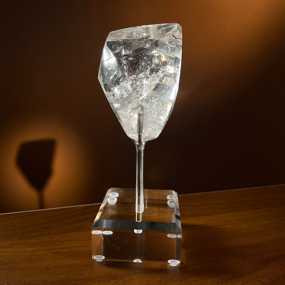 Alt Text: A highly transparent natural clear quartz freeform crystal resting on a modern acrylic display stand against a dark background.
