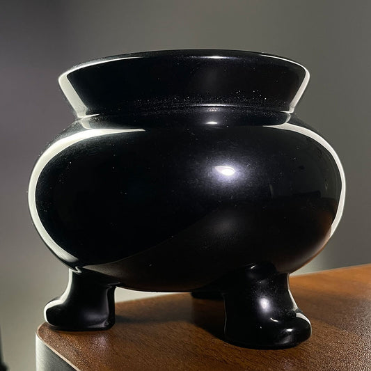  A large, highly polished black obsidian crystal cauldron resting on a wooden table, featuring a sleek three-legged tripod design.