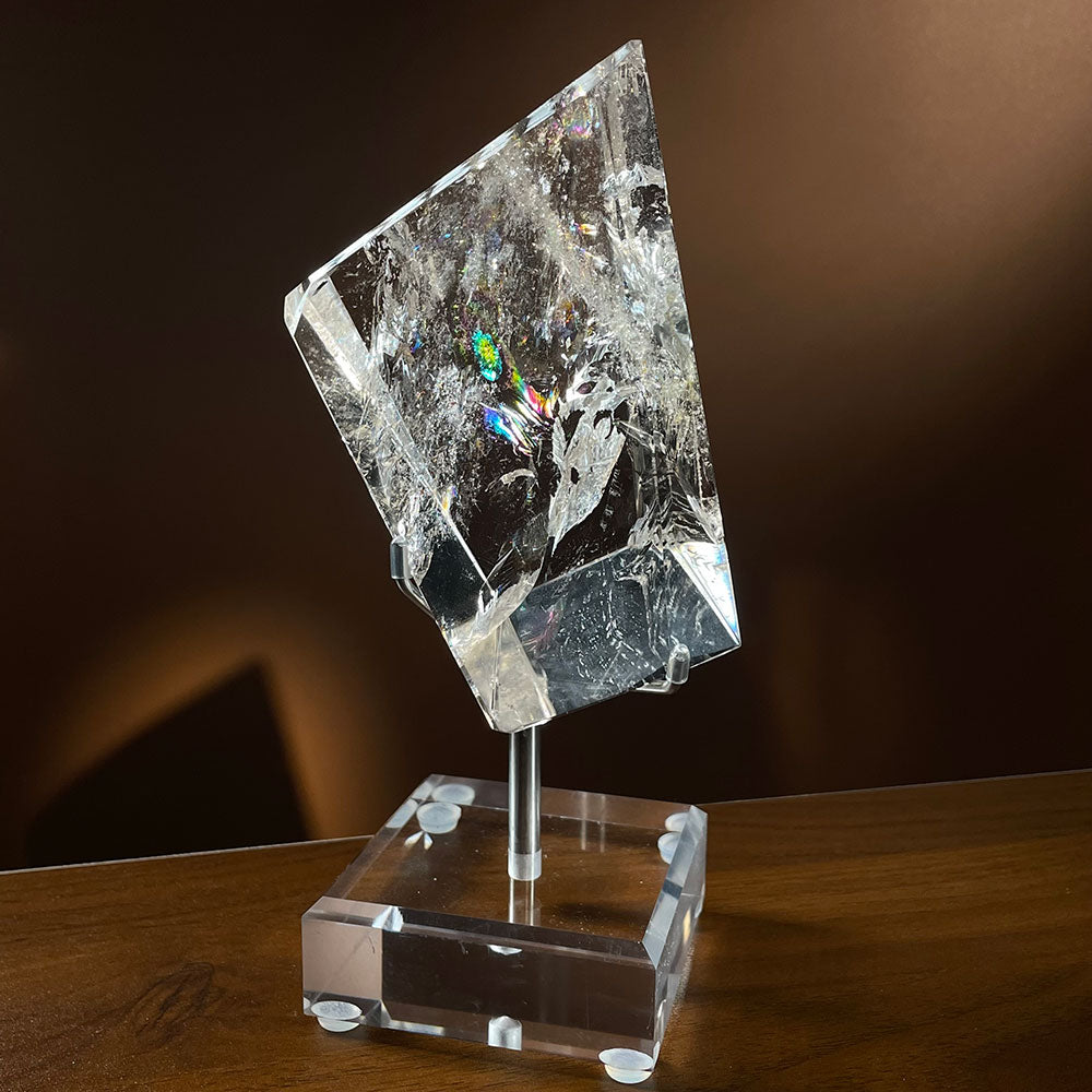 A large 1.03kg water-clear natural quartz polished freeform resting on a clear acrylic stand against a warm brown background.