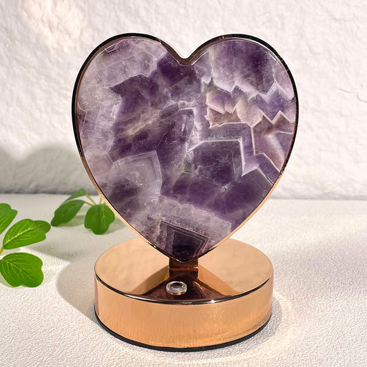  A natural dream amethyst crystal slice carved into a heart, framed in a rose gold metal base, resting on a white surface.
Image B: