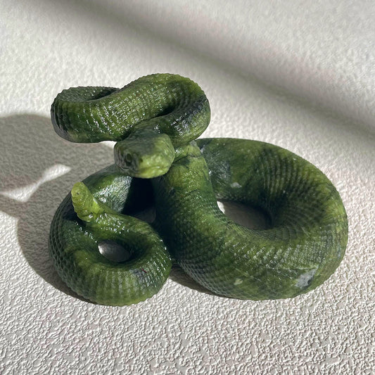 A highly detailed, hand-carved green serpentine crystal coiled rattlesnake figurine with a raised tail, resting on a white surface.
