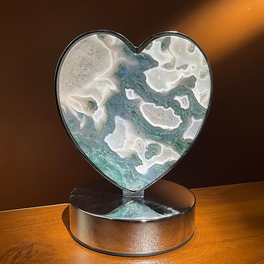 A natural green and white moss agate crystal slice carved into a heart, framed in silver metal on a base, shown unlit on a wooden table.